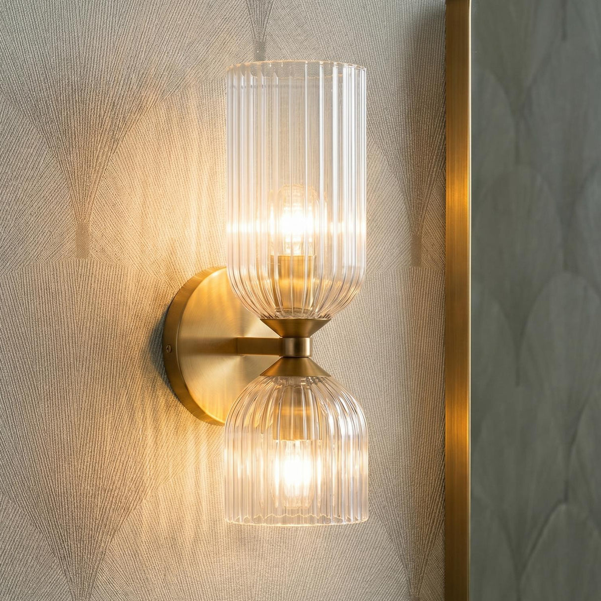 A contemporary bathroom with an Elle Wall Sconce, Brushed Brass and Clear Glass with ribbed shades, mounted beside a gold framed mirror on fan patterned wallpaper.