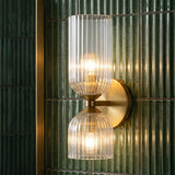 A modern bathroom featuring the Elle Wall Sconce, Brushed Brass and Clear Glass, mounted on vertical green fluted tile beside a gold mirror frame.