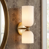 A contemporary bathroom with an Elle Wall Sconce, Brushed Brass and Milk Glass featuring ribbed shades, mounted on textured wallpaper beside a round gold mirror and window.