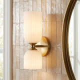 A transitional bathroom with an Elle Wall Sconce, Brushed Brass and Milk Glass mounted on a textured beige tile wall beside a round gold mirror.