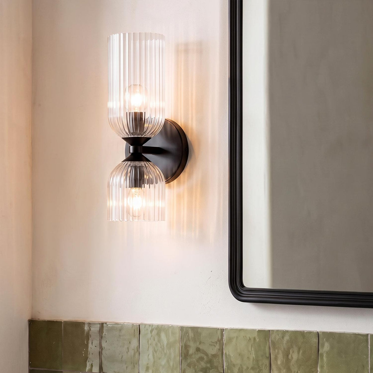 A contemporary bathroom with the Elle Wall Sconce, Matte Black and Clear Glass mounted on a white wall beside a black mirror above sage green tiles.