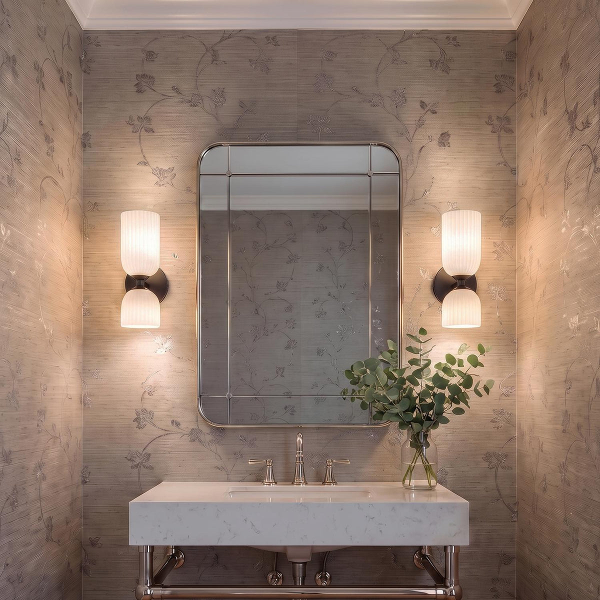 A transitional bathroom with two matte black and ribbed milk glass wall sconces flanking a silver-framed mirror over a white marble vanity with floral wallpaper.