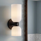 A modern bathroom features the Elle Wall Sconce, Matte Black and Milk Glass, mounted next to a decorative mirror above a marble backsplash.