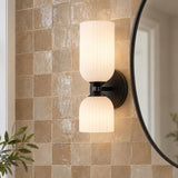 A transitional bathroom features the Elle Wall Sconce, Matte Black and Milk Glass mounted beside a round black mirror on a beige glazed zellige tile wall.