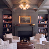 A traditional living room featuring a brushed brass semi-flush mount with ribbed glass shades on a dark gray coffered ceiling above four plaid armchairs and built-in bookshelves.