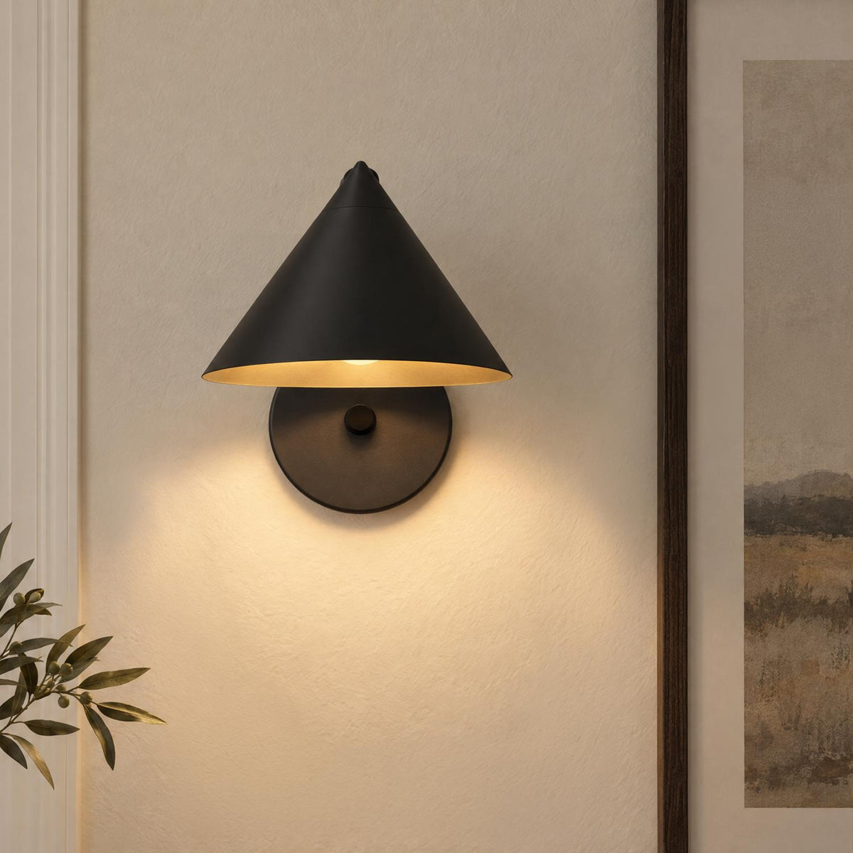 A modern living room displays the Juno Wall Sconce, Matte Black with a conical shade mounted on a beige wall beside framed landscape artwork.