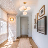 A transitional foyer featuring the Flynn 5 Light Pendant 18", Brushed Brass with Opal Glass along the ceiling above a scalloped jute rug and patterned wallpaper.