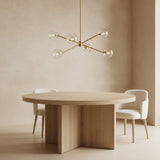 Flynn 6 Light Chandelier 37" wide, in Brushed Brass with Clear Glass, shown in a modern room with a round wooden dining table and ivory boucle chairs.