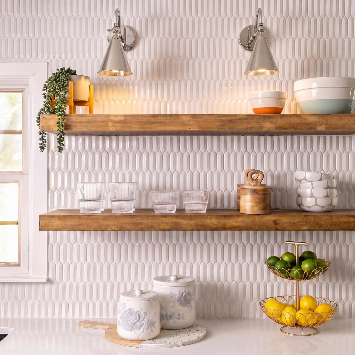 A transitional kitchen with two polished nickel Faye Adjustable Wall Sconces mounted above wood floating shelves and a white textured picket tile backsplash.