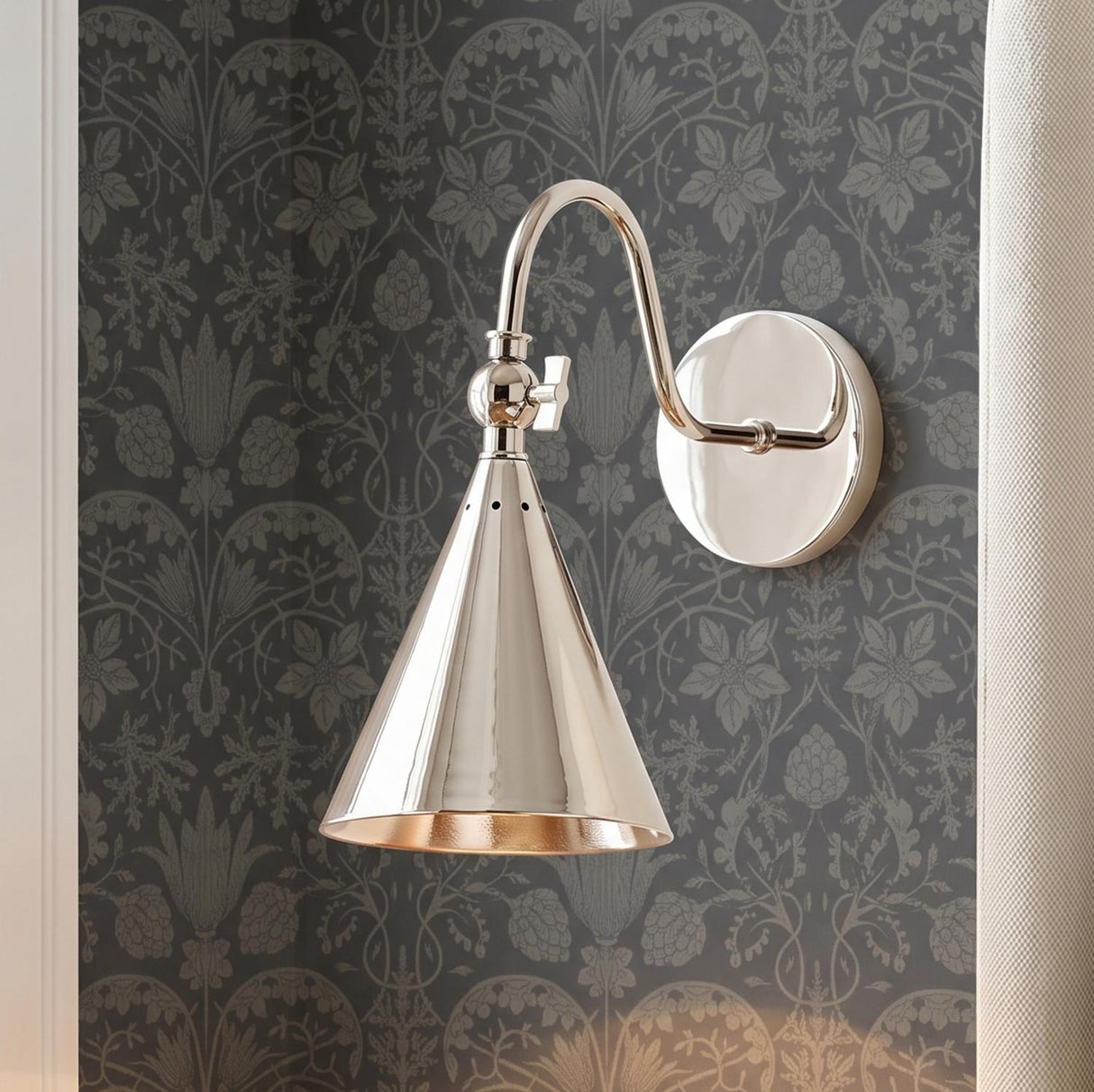 A traditional bedroom with a polished nickel Faye Adjustable Wall Sconce with a cone shade mounted on dark grey botanical wallpaper beside a beige fabric curtain.