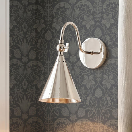 A traditional bedroom with a polished nickel Faye Adjustable Wall Sconce with a cone shade mounted on dark grey botanical wallpaper beside a beige fabric curtain.