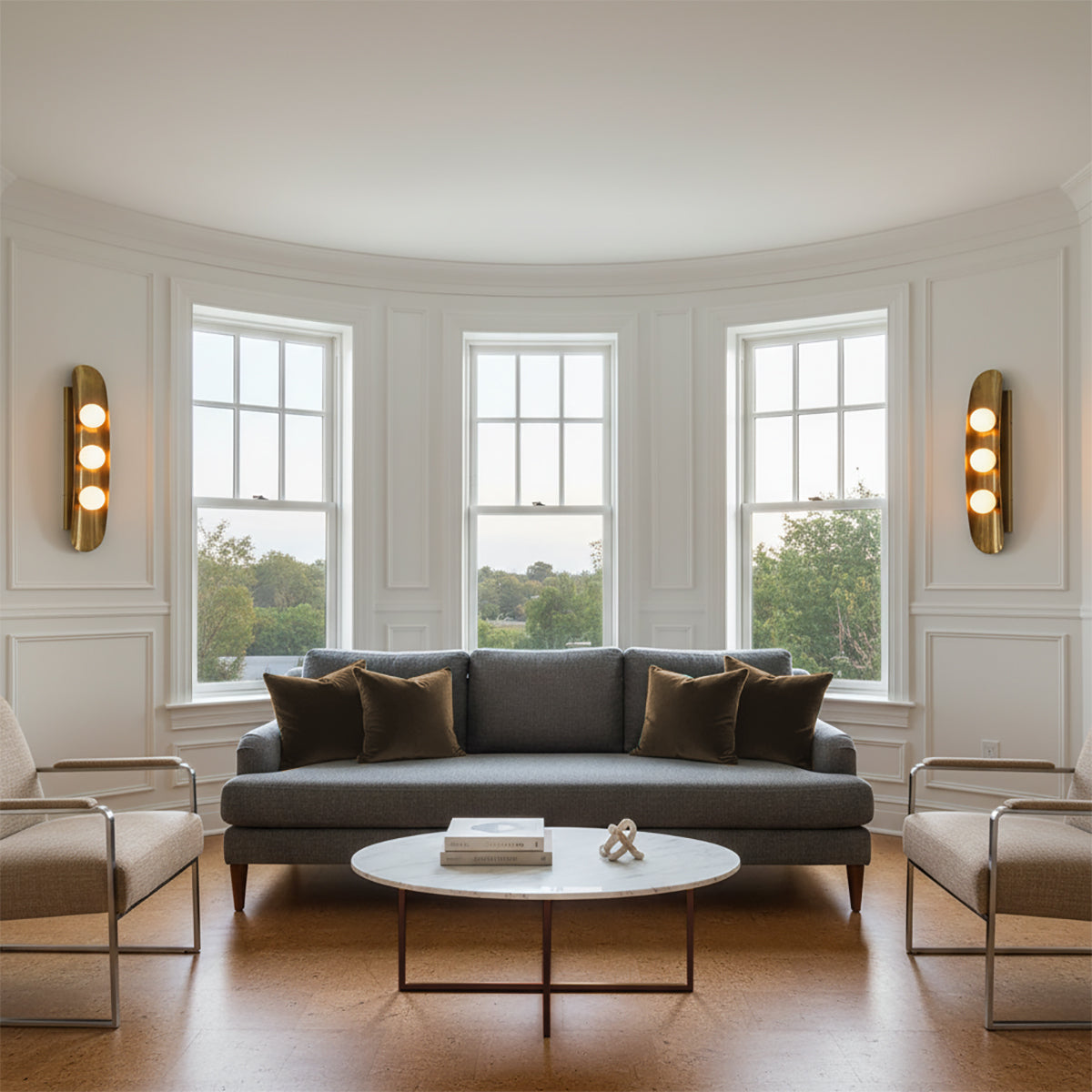 Jen Wall Sconce 24, Brass with Black Accents - Two brass wall sconces with three glowing orbs each flank three tall windows in a living room, with a gray sofa and coffee table in front of the windows.