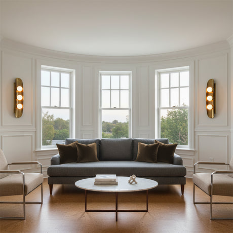 Jen Wall Sconce 24, Brass with Black Accents - Two brass wall sconces with three glowing orbs each flank three tall windows in a living room, with a gray sofa and coffee table in front of the windows.