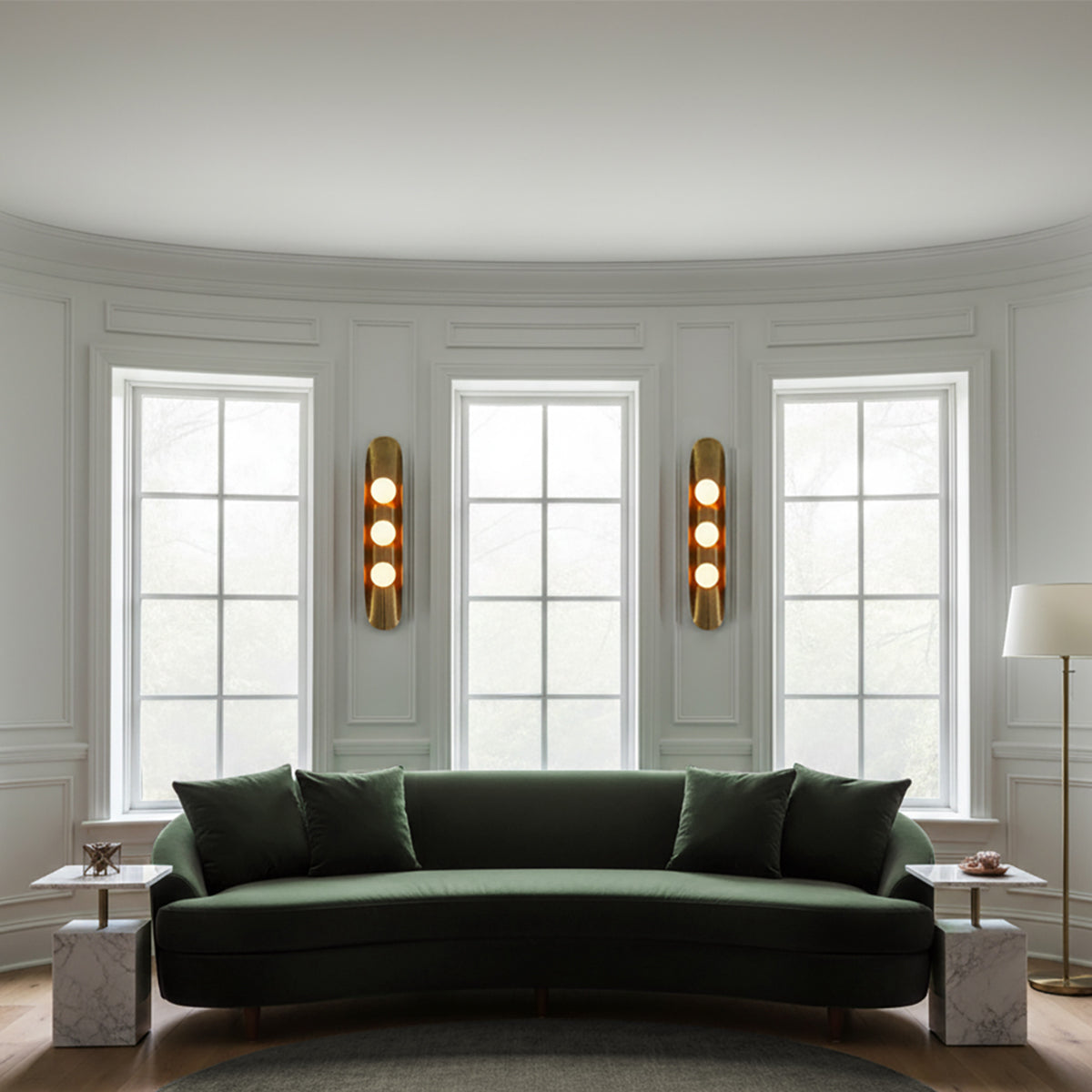Jen Wall Sconce 24, Brass with Black Accents - Two brass wall sconces with three glowing orbs are mounted on a white wall, flanking three large windows with a green sofa in front.