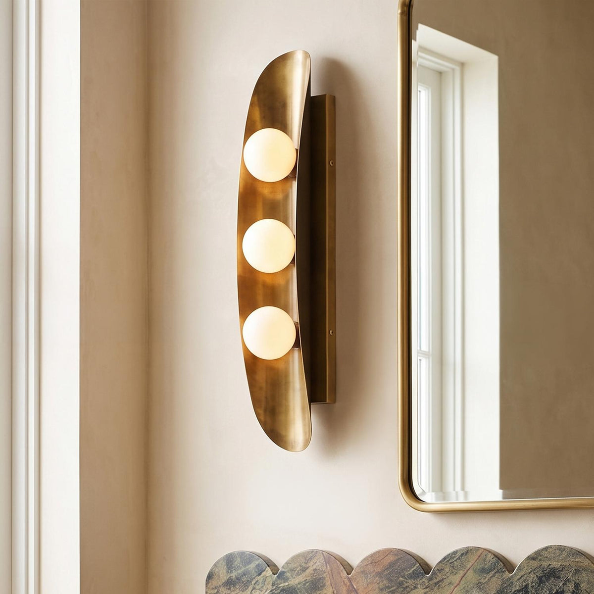 A contemporary bathroom features a brushed brass wall sconce with three frosted glass globes mounted beside a curved brass mirror above a scalloped marble backsplash.