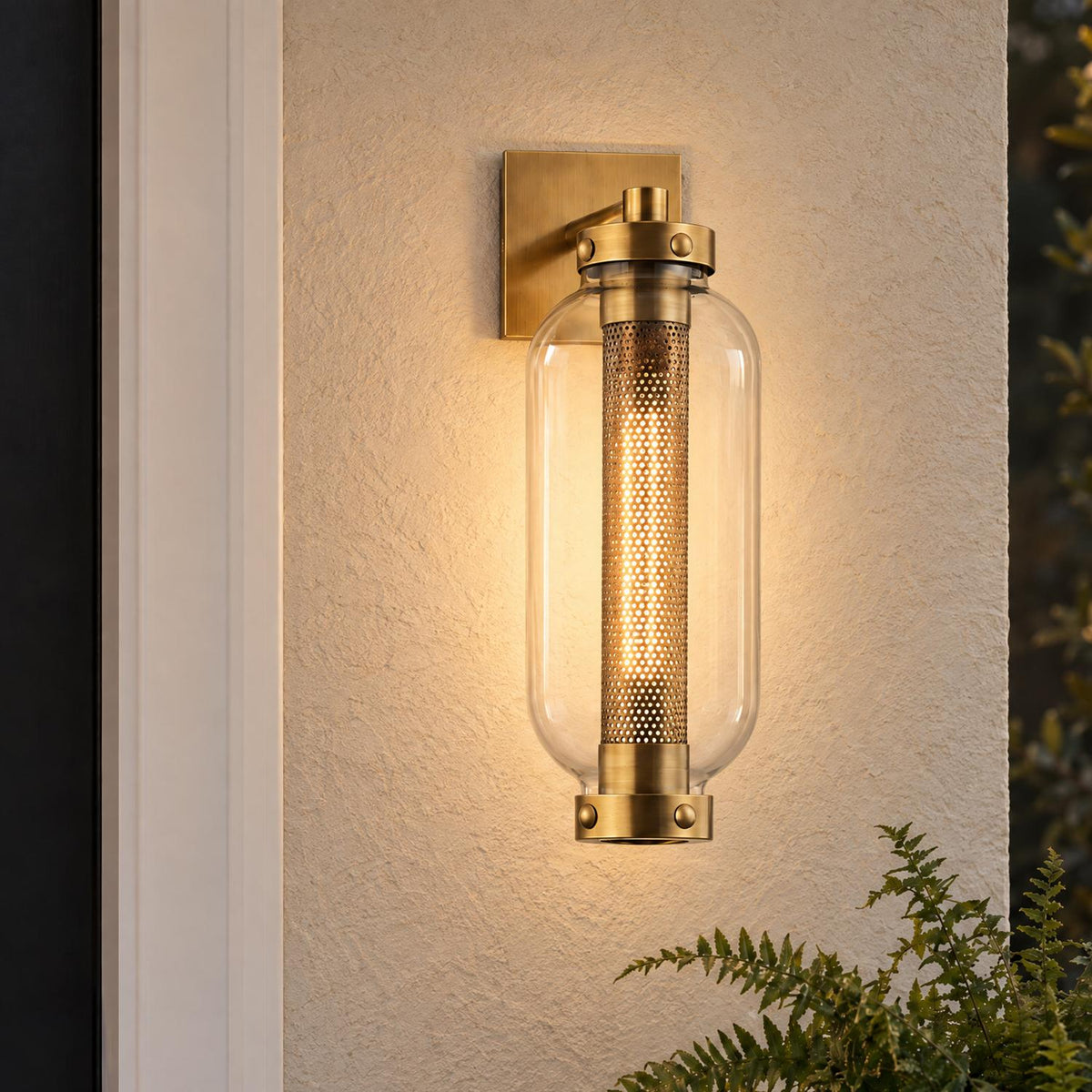 A modern home exterior featuring the glowing Bennett Outdoor Wall Light, Medium 19" Vintage Brass mounted on beige stucco beside a dark door with green fern leaves.