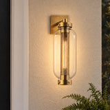 A modern home exterior featuring the glowing Bennett Outdoor Wall Light, Medium 19" Vintage Brass mounted on beige stucco beside a dark door with green fern leaves.
