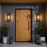 A modern entry featuring a light wood plank door flanked by bronze wall lanterns with vintage brass accents on smooth gray walls at night beside potted boxwoods.