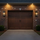 Orin Outdoor Wall Light, Large 24" Bronze with Vintage Brass Accents - Two rectangular outdoor wall lanterns with glass panels and visible candle-style lights flank a dark wood double garage door set in a brick wall, illuminated by upward-facing exterior lights.