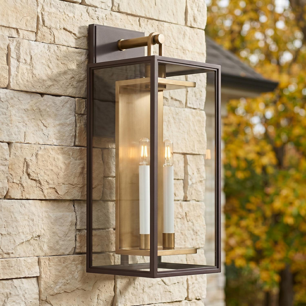 A transitional home exterior featuring the Orin Outdoor Wall Light in bronze with vintage brass accents mounted on a textured cream stone wall with autumn foliage background.