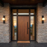 A modern home entry with two Gus Outdoor Wall Light, Large 24" Textured Black fixtures flanking a wood front door on stacked stone at dusk.
