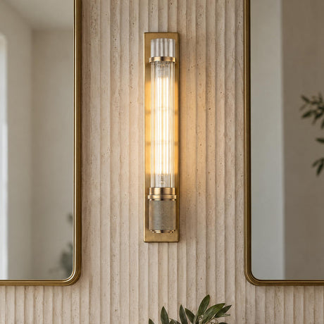 A modern bathroom vanity featuring the Zeke Wall Sconce 16", Aged Brass with fluted glass, mounted on a beige ribbed stone wall between brass framed mirrors.