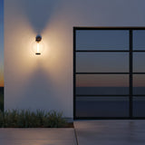 Rogers Outdoor Wall Light, 18" Bronze - A round, clear glass outdoor wall light with a bronze fixture is mounted on a textured white wall, casting a warm glow on the wall and illuminating lush green grass below. A large, dark-paned window is visible to the right.