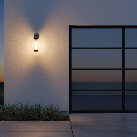 Rogers Outdoor Wall Light, 18" Bronze - A round, clear glass outdoor wall light with a bronze fixture is mounted on a textured white wall, casting a warm glow on the wall and illuminating lush green grass below. A large, dark-paned window is visible to the right.