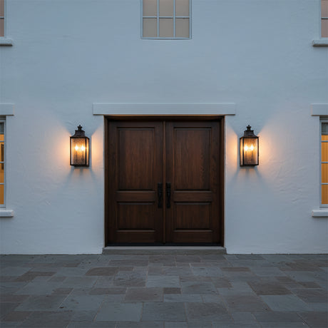 Bedford Outdoor Wall Light, Medium 21" Antique Black Iron - Two lanterns illuminate a dark wood double door entrance with stone paving outside a white stucco building.