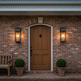 Monroe Outdoor Wall Light, Medium 19" Forged Iron with Vintage Brass Accents - Two lanterns flank a wooden door on a brick wall, with plants and a bench.