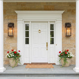 A traditional stone entry with large Monroe Outdoor Wall Light lanterns in forged iron with vintage brass accents flanking a white door with potted geraniums at dusk.