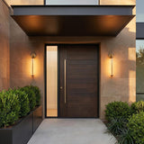 A modern home entry with Marcy LED Outdoor Wall Light, 21" Vintage Brass sconces flanking a dark wood door on stone panels at twilight with boxwood hedges.