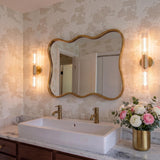 A transitional bathroom featuring two aged brass wall sconces with ribbed glass flanking a wavy gold mirror above a white vessel sink and marble countertop.