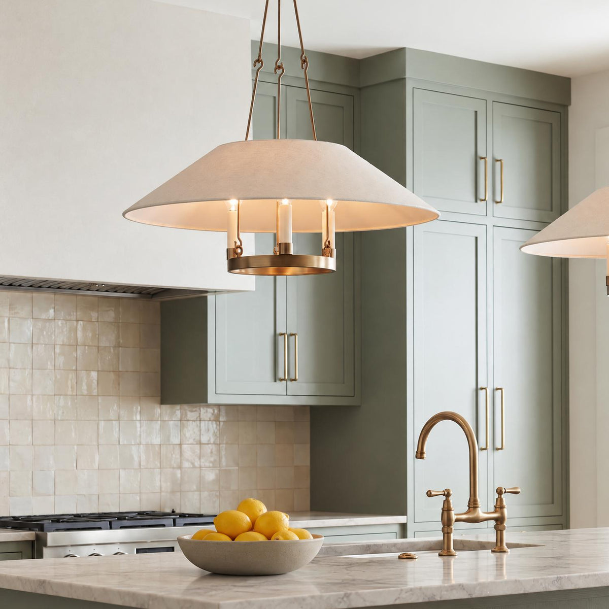 A transitional kitchen features the Dawson Pendant 25", Patina Brass with Linen Shade over a marble island with sage green cabinets and brass faucet.