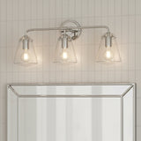 Addison 3 Light Vanity Light, Polished Nickel - Three clear glass cone-shaped shades with exposed Edison bulbs mounted on a polished nickel horizontal bar against a white tiled wall above a mirrored surface.