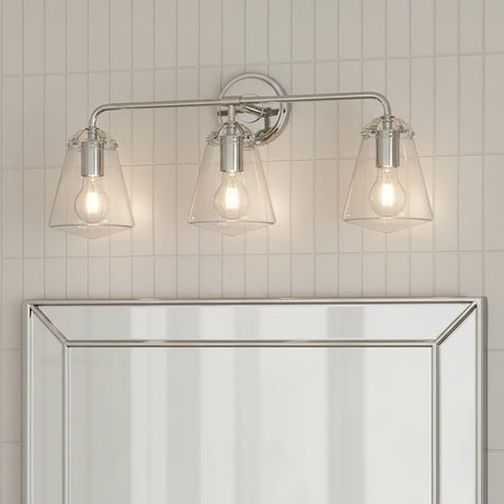 Addison 3 Light Vanity Light, Polished Nickel - Three clear glass cone-shaped shades with exposed Edison bulbs mounted on a polished nickel horizontal bar against a white tiled wall above a mirrored surface.