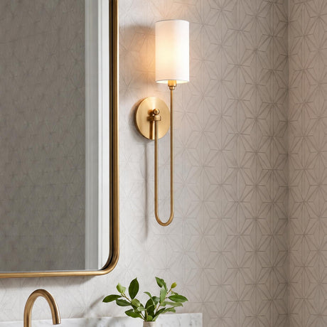 A transitional bathroom featuring an aged brass wall sconce with a white fabric shade mounted on geometric wallpaper beside a brass mirror above a marble countertop.