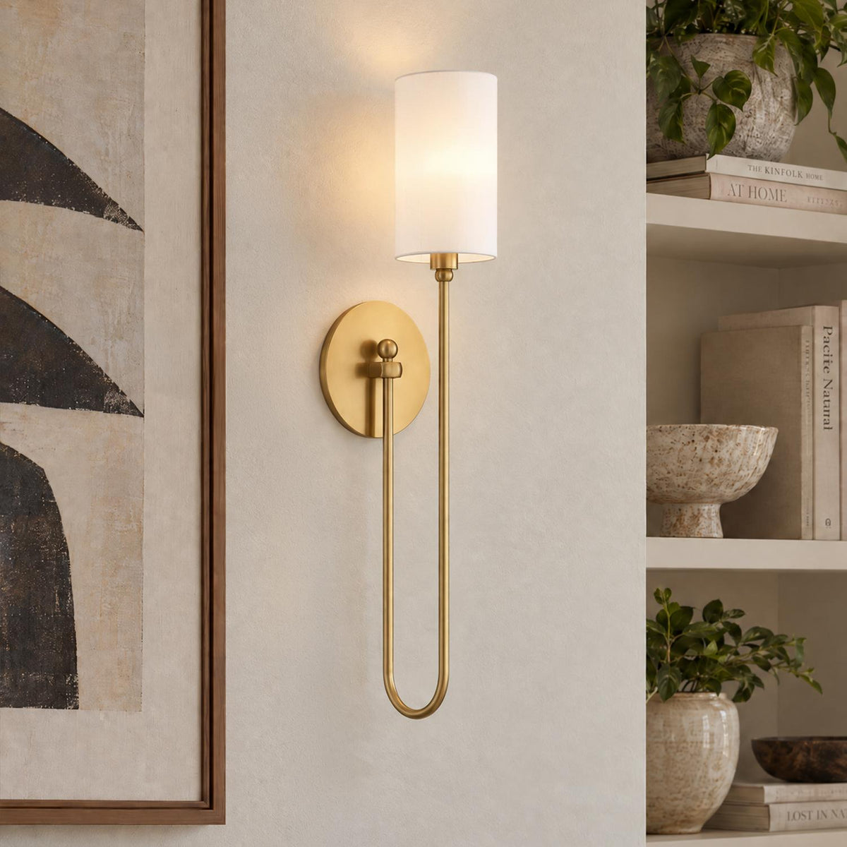 A transitional living room features the Briar Wall Sconce 24", Aged Brass with a white shade mounted on a textured wall beside built-in decorative shelves.