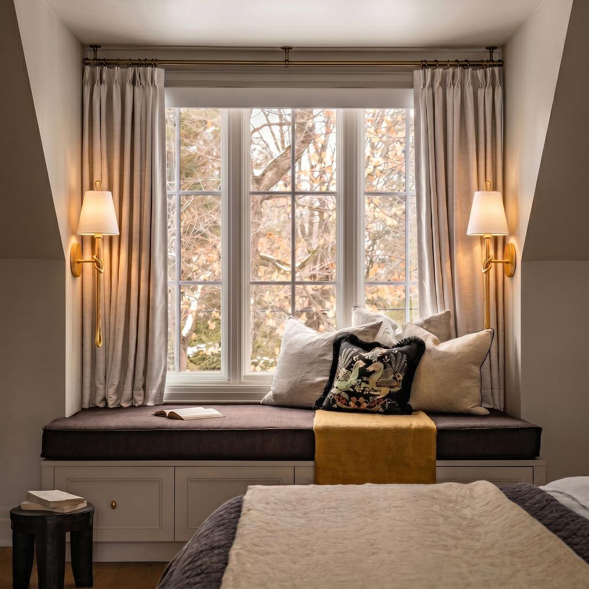 A traditional bedroom with two gold leaf Archer Wall Sconce 24" fixtures featuring white linen shades flanking a built-in window seat under a sloped ceiling.