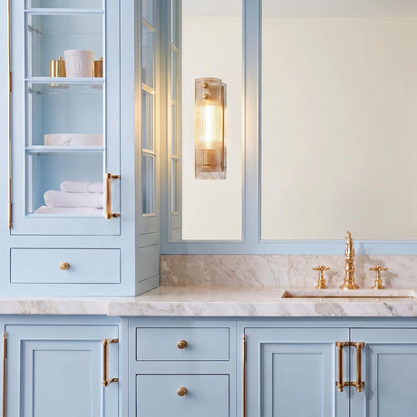 A transitional bathroom featuring the Jensen 1 Light Wall Sconce 14", Aged Brass with ribbed glass beside a mirror above light blue cabinetry and marble countertop.