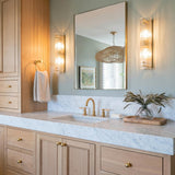 A transitional bathroom with Jensen 2 Light Wall Sconce 23", Aged Brass fixtures featuring ribbed glass flanking a mirror over a marble countertop and light wood vanity.