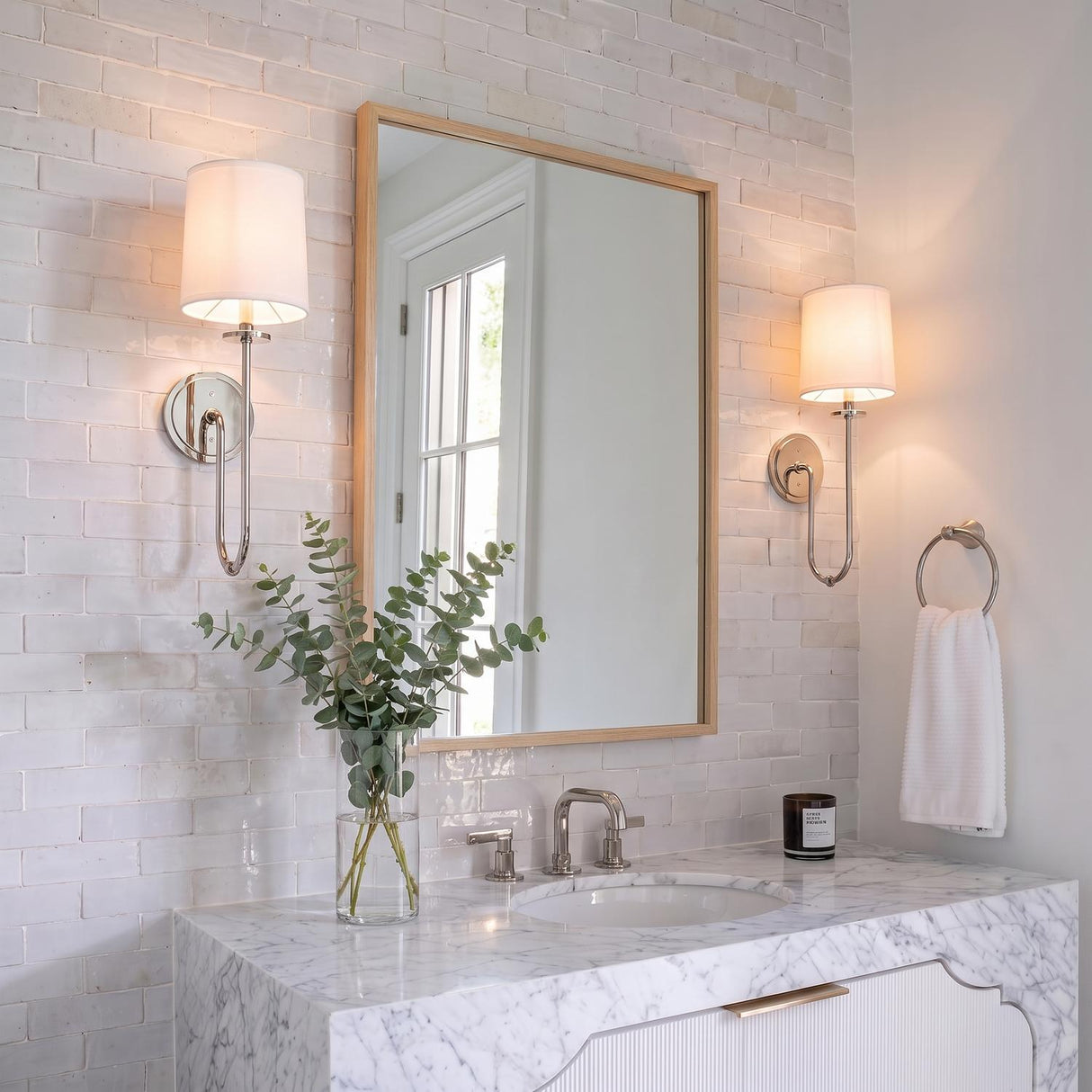 A transitional bathroom features two Hayden Wall Sconce 21", Polished Nickel fixtures with linen shades flanking a wood-framed mirror above a white marble waterfall vanity.
