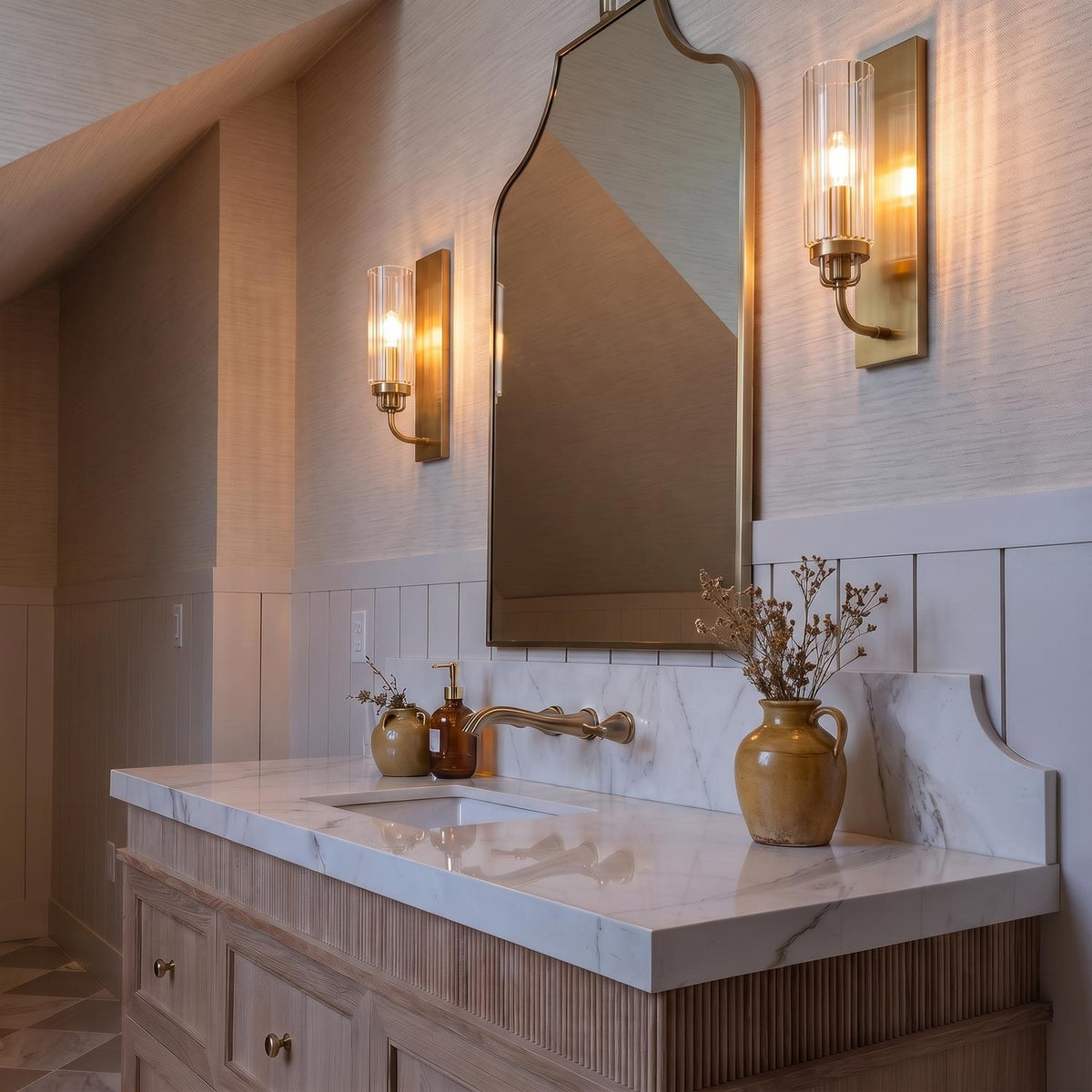A transitional bathroom with two Mercer Wall Sconce 14", Aged Brass sconces flanking an arched mirror above a white marble vanity under a sloped ceiling.