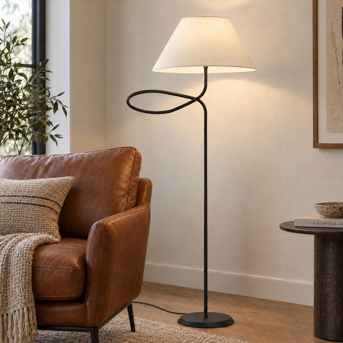 A cozy modern living room features the Odin Floor Lamp, Forged Iron with a looped matte black stem beside a cognac leather armchair.