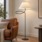 A cozy modern living room features the Odin Floor Lamp, Forged Iron with a looped matte black stem beside a cognac leather armchair.