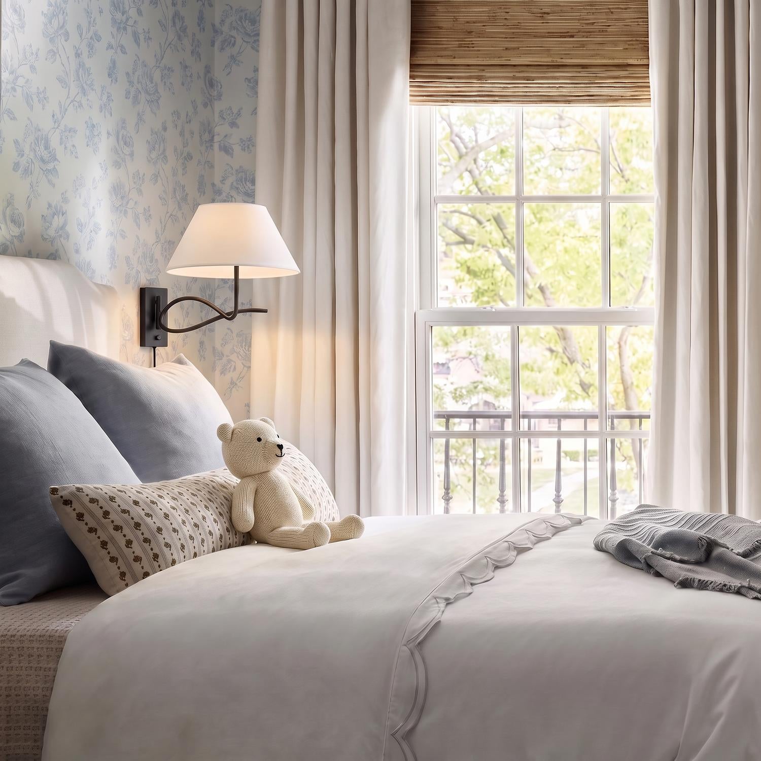 A traditional bedroom features an Odin Plug-In Wall Sconce, Forged Iron with a white linen shade mounted on blue floral wallpaper beside a white scalloped duvet.