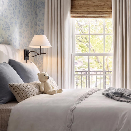 A traditional bedroom features an Odin Plug-In Wall Sconce, Forged Iron with a white linen shade mounted on blue floral wallpaper beside a white scalloped duvet.