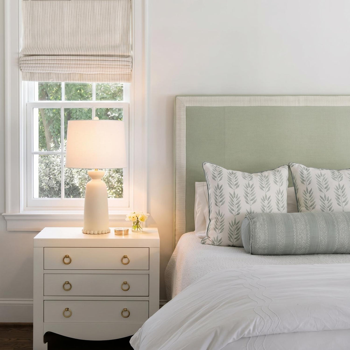 A traditional bedroom with the Bonnie Ceramic Table Lamp, Antique Ivory featuring a linen shade on a white nightstand beside a sage green headboard.