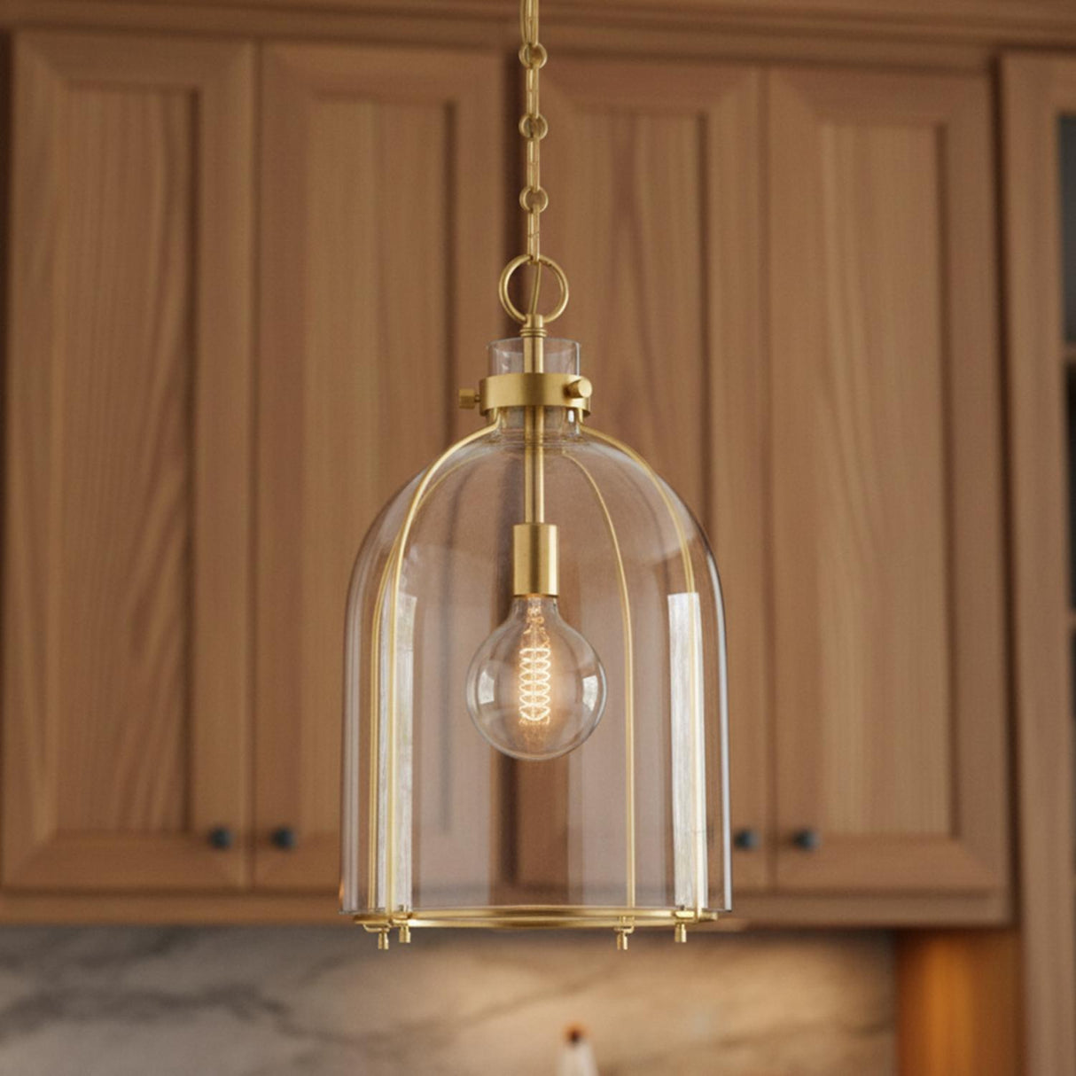 A transitional kitchen features the Lana Pendant 16", Aged Brass and Clear Glass hanging over the island against light wood cabinets and a marble backsplash.