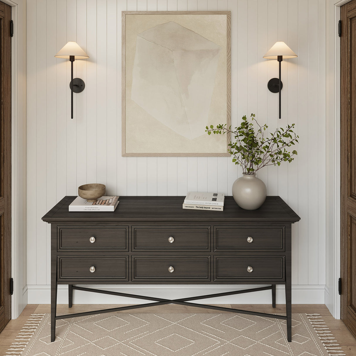 Two Scarlett 26" tall Wall Sconces, in Forged Iron finish, are above a dark wood console table with decorative items in a room with white paneled walls and wooden flooring.